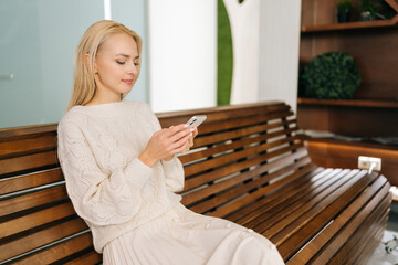 Medium shot of young blonde woman sitting on wooden bench using smartphone looking at cellphone screen enjoying using mobile apps. Happy female checking social media holding smartphone.