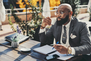 A black balding corporate man with a beard sitting in an outdoor cafe, dressed in a fancy suit, polka-dot tie, and a white vest, reviews his notes with a pen, and while taking a sip of his coffee - obrazy, fototapety, plakaty