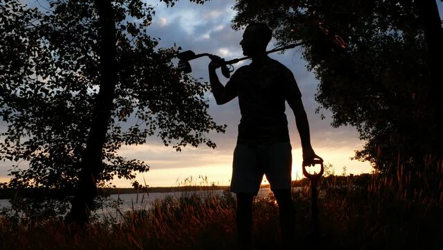 The silhouette of a man at sunset who walks with a metal detector in search of precious metals. Outdoors among the vegetation on the background.
