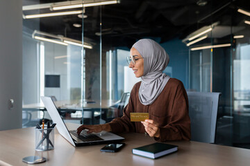 Successful muslim woman in hijab at work inside modern office, business woman with bank credit card and laptop makes online shopping in online store, satisfied and smiling woman at workplace.