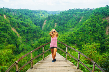 Fototapeta na wymiar Family vacation lifestyle. Young woman stand on edge of overhanging balcony on high cliff. Happy girl looking at stunning tropical jungle view. Tukad Melangit is popular travel destination in Bali