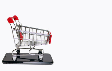 A small shopping cart stands on a mobile phone on a white background. The concept of online...