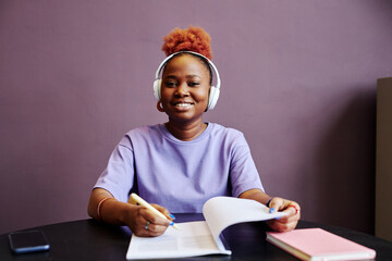 Minimal portrait of smiling black woman wearing headphones and taking notes in minimal purple...