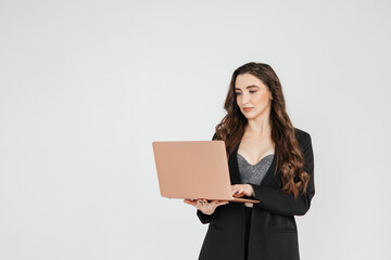 Curly adult business lady in elegant black suit standing while working on laptop against gray background