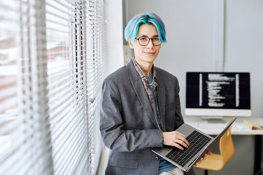 Portrait of Asian young man with colored blue hair looking at camera and holding laptop by window