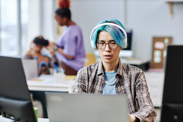 Portrait of young Asian man with blue hair using computer in office and wearing headphones