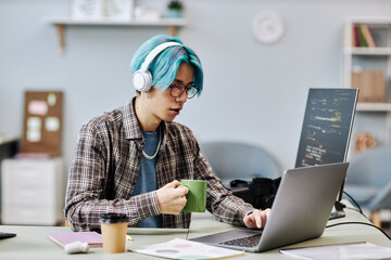 Portrait of young man with blue hair using computer in office and wearing headphones while writing...