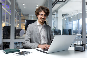 Fototapeta premium Portrait of young successful businessman financier, man inside office at workplace sitting using laptop smiling and looking at camera, happy with achievement freelancer.