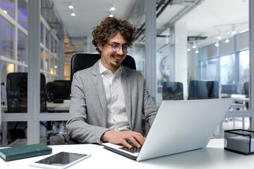 Fototapeta premium Cheerful and successful businessman inside office working with laptop, bearded man smiling and typing on laptop keyboard, programmer coding software in modern room.