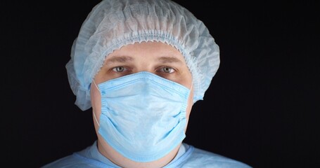 The surgeon looks at the camera in a blue uniform, surgical gown, mask and cap. On a black isolated background, a male surgeon looks at the camera. Portrait, close-up.