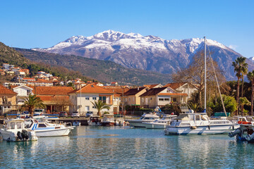 Fototapeta na wymiar Beautiful winter Mediterranean landscape. Montenegro, Tivat city. View of Marina Kalimanj on sunny day. Fishing boats in harbor. Snow-capped Lovcen mountains
