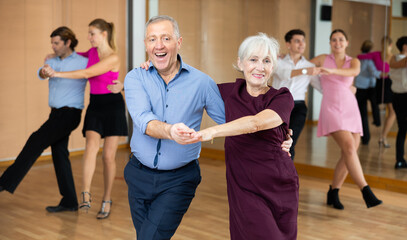 Cheerful elderly woman and man enjoying active dancing in pair during group training in dance studio, practicing playful jitterbug moves - obrazy, fototapety, plakaty