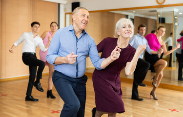 Positive aged man and woman dancing energetic upbeat jive as couple during group training in dance studio..