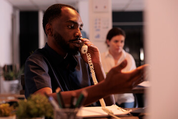Project manager discussing company report with colleague on landline phone, employee answering call in office late. African american man having telephone communication in coworking space