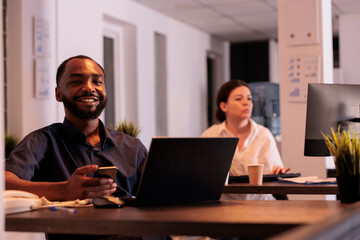 Smiling african american man working on report on laptop, using smartphone software in coworking space portrait. Happy project manager looking at camera at workplace desk