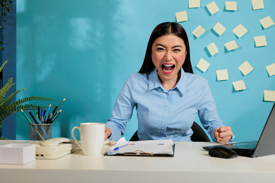 Overworked female corporate employee expressing desperation and stress by shouting loudly at the camera. Professional woman with negative feelings due to exaggerated workload.