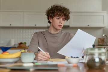 Fototapeta na wymiar Close up of young curly haired handsome man wearing beige t-shirt sitting at a kitchen table surrounded by comforts of domestic life making notes reviewing papers while having breakfast in kitchen.