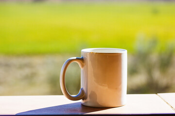 White cup mug of tea or coffee standing on windowsill against green grass outside window on sunny warm day. A morning drink without people on a nature in summer. Selective focus. Relax, lounge concept