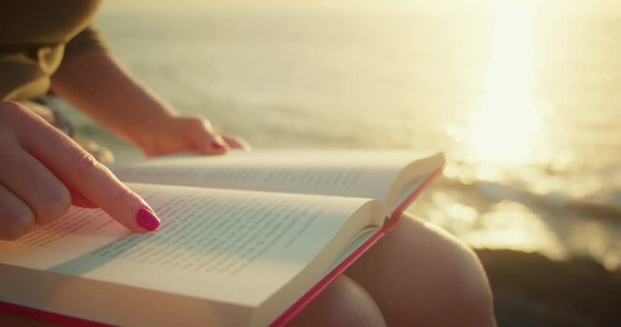 Girl self-learning by reading a book outdoors. Ocean waves on the beach at sunset. Close-up of woman hands. Finger points to printed text.