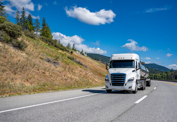 Long haul low cab profile white big rig semi truck transporting liquid cargo in tank semi trailer driving on the winding mountain road
