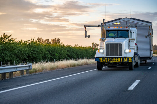 Powerful white big rig classic semi truck tractor with oversize load sign on the bumper transporting oversized manufactured house driving on the road at twilight evening