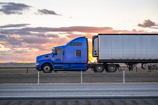 Side view of the blue classic bonnet big rig semi truck with extended cab transporting cargo in refrigerated semi trailer driving on the highway road at sunset twilight