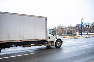 White day cab middle rig semi truck with long box trailer transporting goods to local customers driving on the slippery winter city road in New England