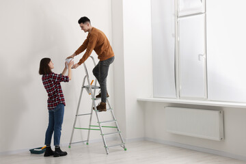 Young woman giving paint can to boyfriend on stepladder indoors. Room renovation