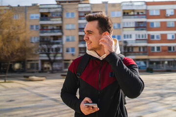 A young guy wearing wireless headphones or earbuds and using his phone outside in the city during the day