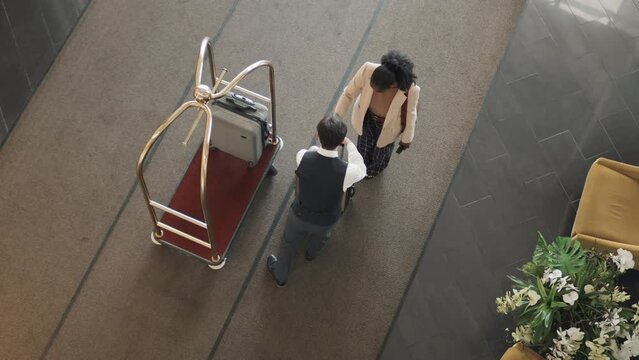 From above view of bellhop wearing uniform working in luxury hotel taking new guests suitcases and carrying them using luggage trolley