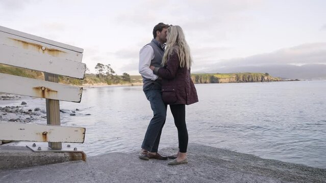 Happy Preppy Romantic Young White Couple during Sunset Golden Hour by the Ocean - wide shot of kissing by water