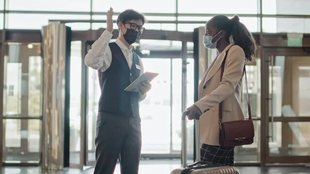 Young Asian concierge wearing mask standing in lobby giving information about hotel to new guest