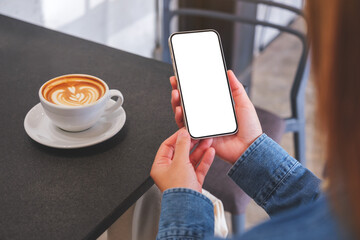 Mockup image of a woman holding mobile phone with blank desktop screen with coffee cup on the table