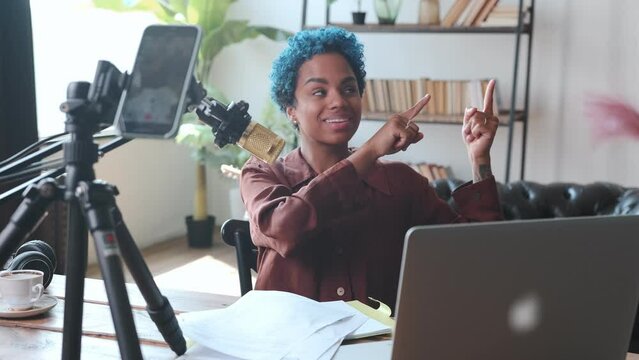 Young happy African American woman sits at table with microphone and mobile phone recording video blog pointing fingers to side to place ad or link to next video located in home studio