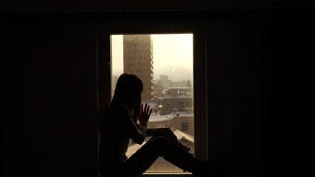 Loneliness Asian woman sitting by the window in bedroom and looking cityscape building covered in snow at winter sunset. Attractive girl resting in hotel room during travel alone on holiday vacation.