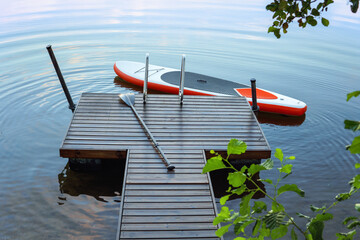 SUP board floating next to the wooden pier on a quiet summer morning. The surface of the lake is...