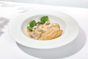 Traditional russian food - beef stroganoff with mushroom sauce and mashed potatoes. Beef stroganoff in white plate on light background with hard shadows. Stew meat with creamy sauce on summer menu.