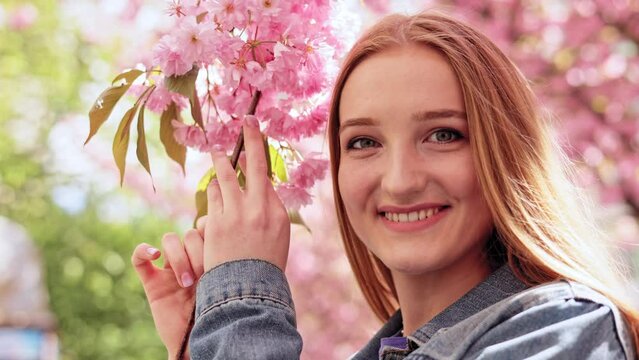 Close up portrait young face woman with beautiful eyes looking and smiling at camera on blurred Sacura tree background in the city street. Pretty Caucasian girl with Cherry blossom trees. Slow motion.