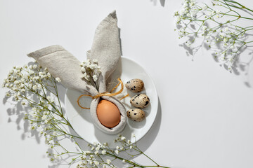Minimalist Easter aesthetic table setting, egg decorated with bunny ears napkin on white plate. Neutral floral background with copy space. Holiday spring template or banner design