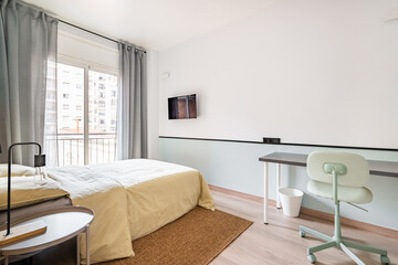 Hotel room with bed for two persons with beige linen. Access to balcony through glass sliding doors. Table against wall with chair for online work. On wall is TV for pleasant evenings watching movie.