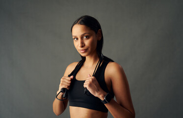 Fototapeta na wymiar Portrait of young biracial woman in sports bra holding jump rope over shoulders in studio
