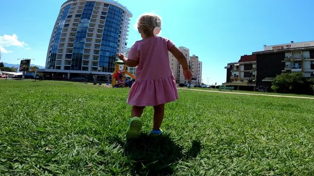 Little girl runs along the green lawn to the playground near the high-rise building