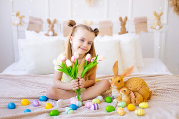 easter, a little girl with painted colored eggs and a rabbit holding spring flowers tulips at home in a bright room preparing for the holiday smiling and having fun