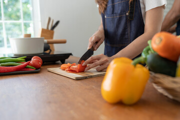 Beautiful Asian woman in kitchen cooking apron preparing various vegetable ingredients and slicing tomatoes in online cooking prep for the health and happiness of our loved ones in their own homes.