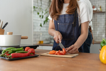 Obraz na płótnie Canvas Beautiful Asian woman in kitchen cooking apron preparing various vegetable ingredients and slicing tomatoes in online cooking prep for the health and happiness of our loved ones in their own homes.