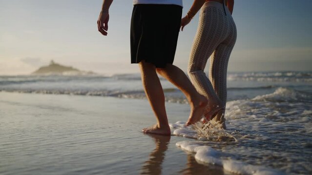 Legs of happy couple holding hands and walking on the beach together enjoying summer side view. Feet of heerful boyfriend and girlfriend relaxing and taking a walk at the seaside at sunrise side shot.