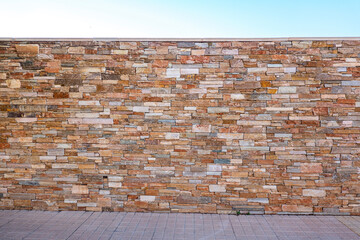 Brown masonry fence of stones, pavement and blue sky. Light brown brick wall background with irregular pattern texture background. Urban decorative stone wall pattern