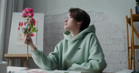 A young female artist admires fresh flowers while looking for inspiration to paint a painting while working in a studio in daylight