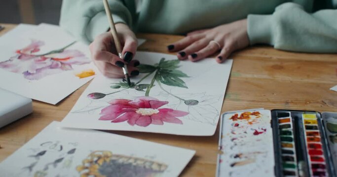 A young artist mixes watercolors with a brush while painting flowers on paper. Video shot in daylight in an art studio