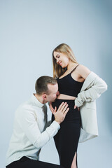 Smiling young man kneeling with his pregnant wife and hugging her belly, isolated on white background. Stylish young people. A man in a shirt and a woman in a black skirt and jacket